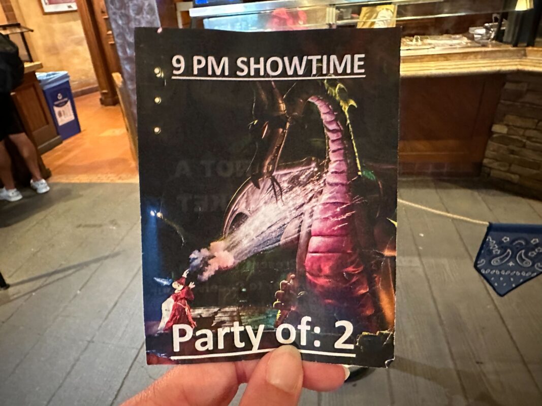 A hand holding a show ticket for a 9 PM performance with the words "Party of: 2" and an image of a dragon and a person on stage.