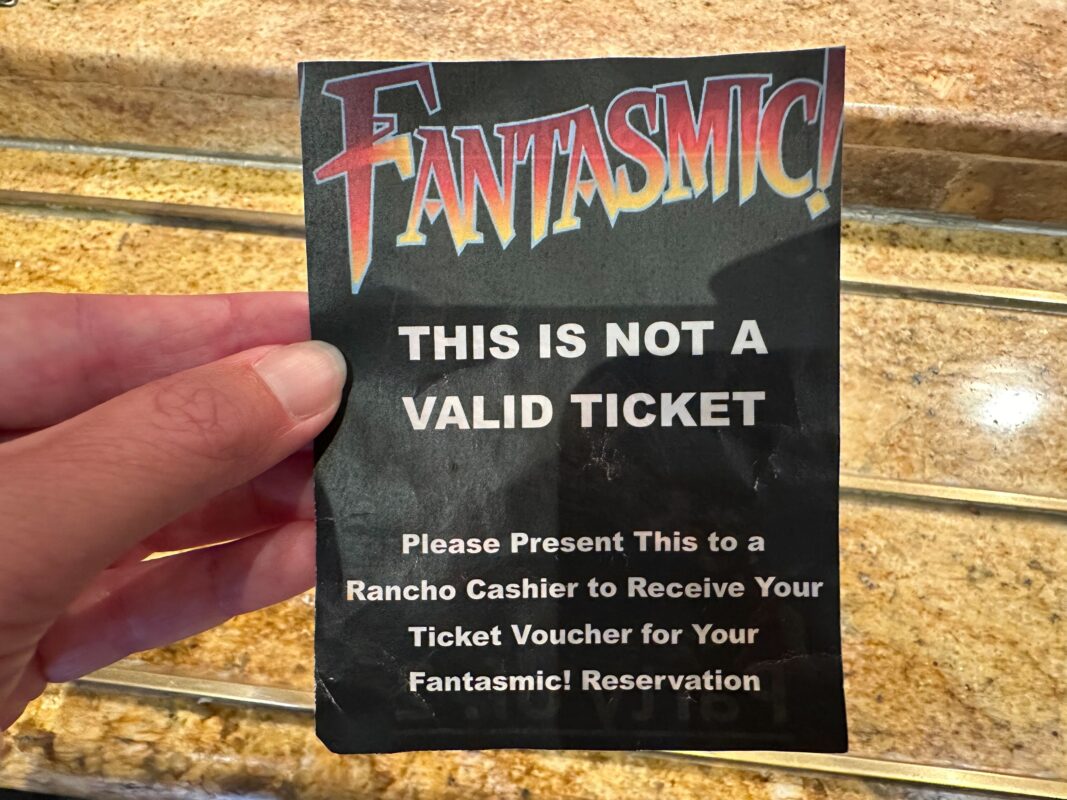 Close-up of a hand holding a paper with "FANTASMIC!" text, informing it is not a valid ticket and instructing to present it to a Rancho cashier for a ticket voucher for a Fantasmic! reservation.