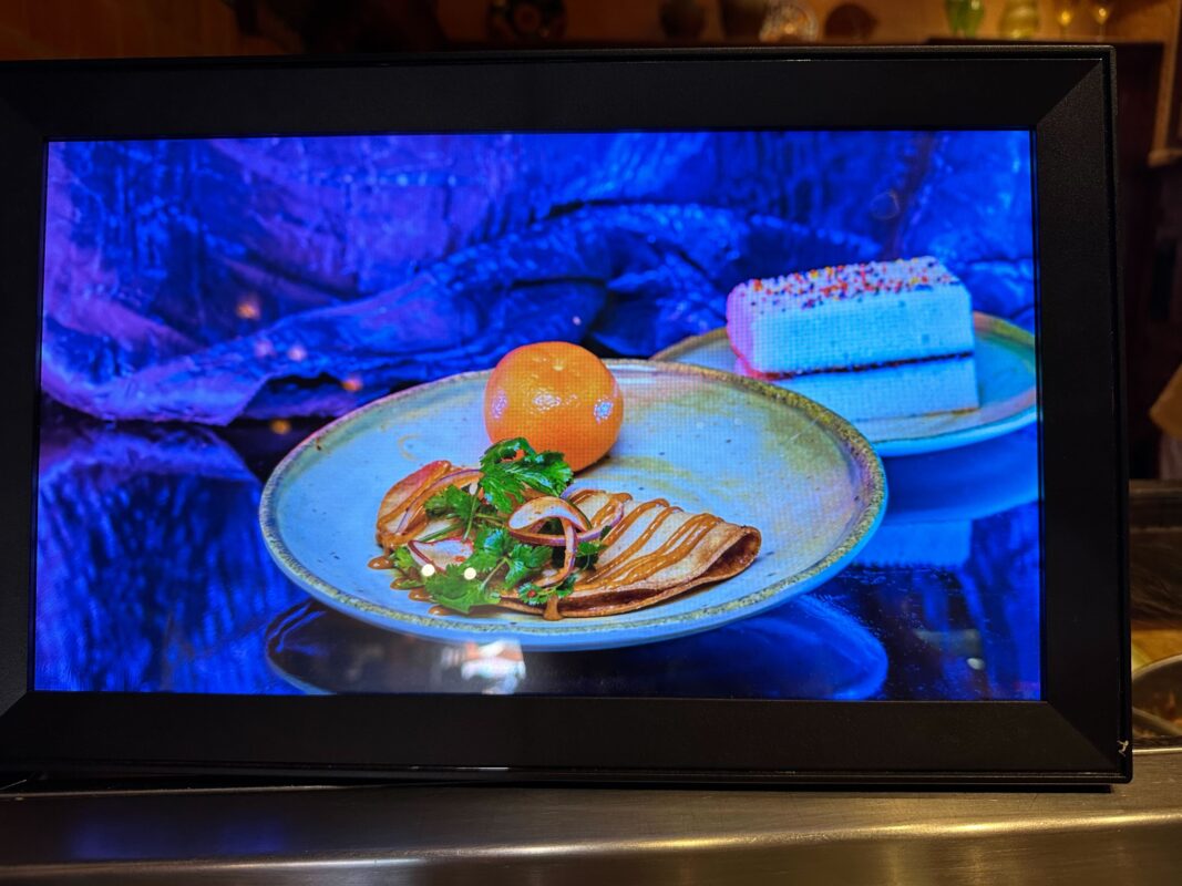 A display showing a plate of food with greens and sauce, a whole orange, and a slice of layered cake with sprinkles on the side.