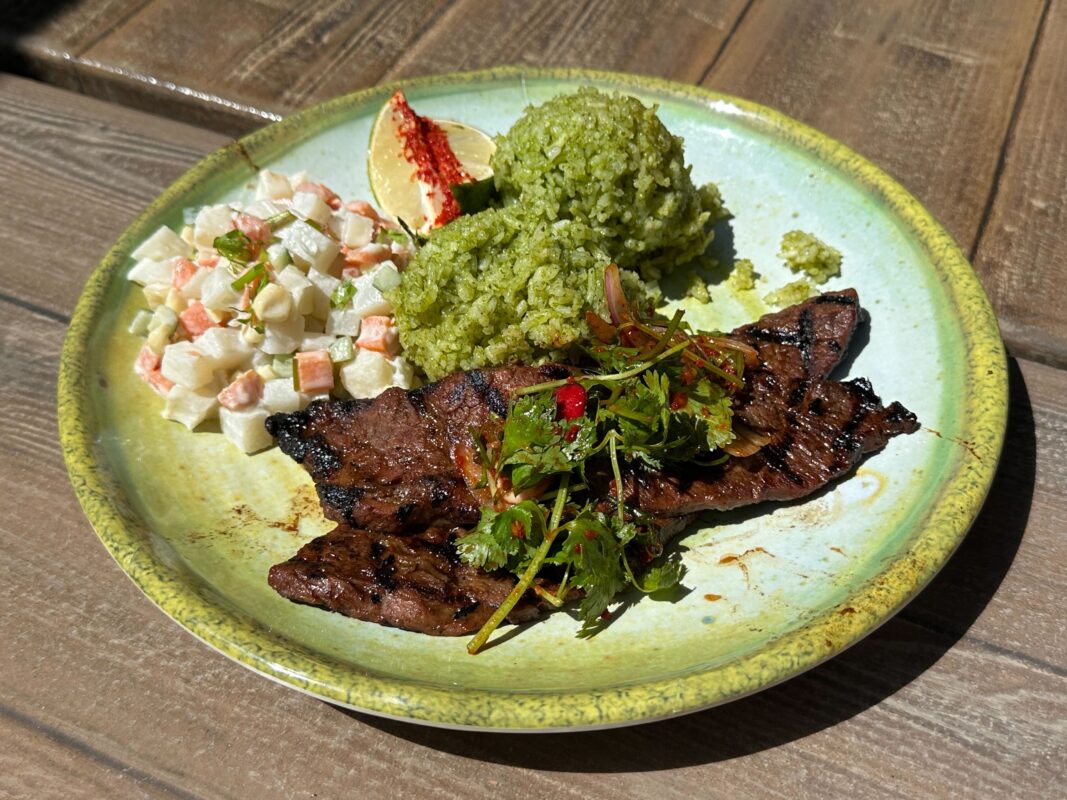 A plate with grilled beef steak, two scoops of green rice, a portion of salad with diced vegetables, and a lime wedge garnish. The food is served on a green ceramic plate on a wooden table.