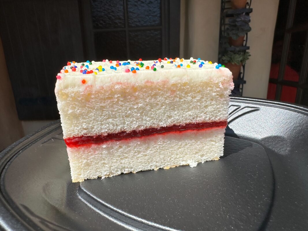 A slice of white cake with frosting and colorful sprinkles on top, featuring a layer of red filling in the middle, placed on a black plate.
