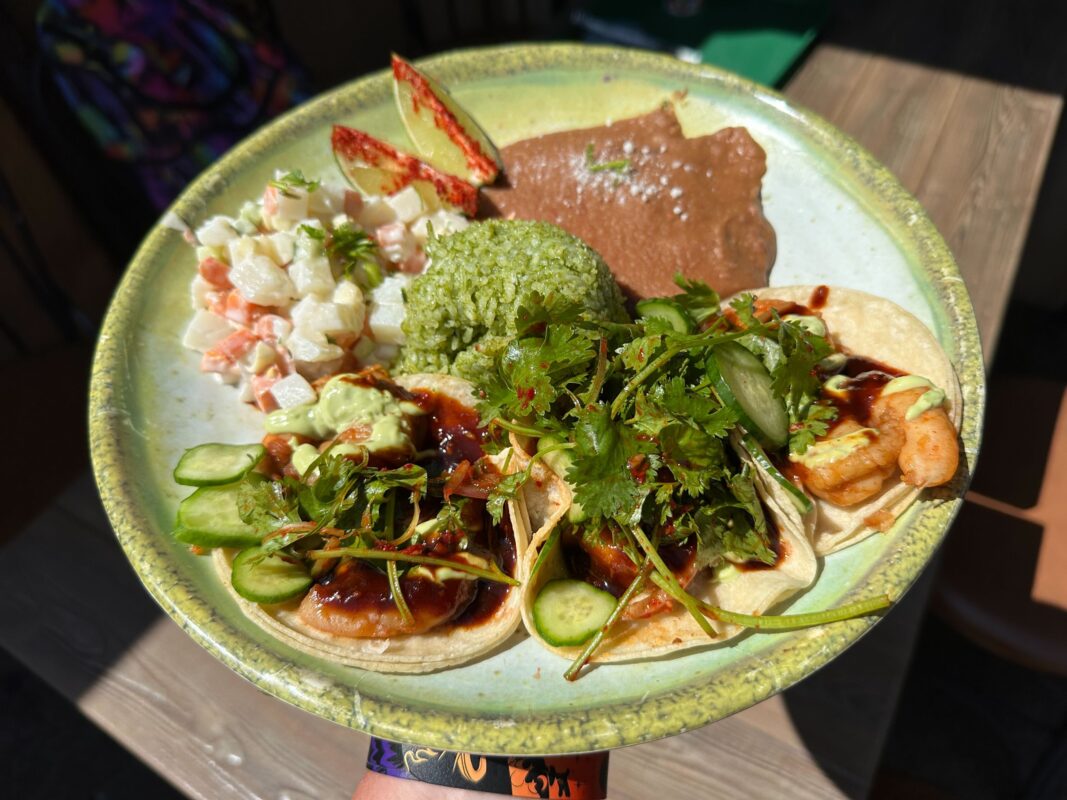 A plate of three tacos topped with green herbs, a side of green rice, refried beans, and a potato salad.