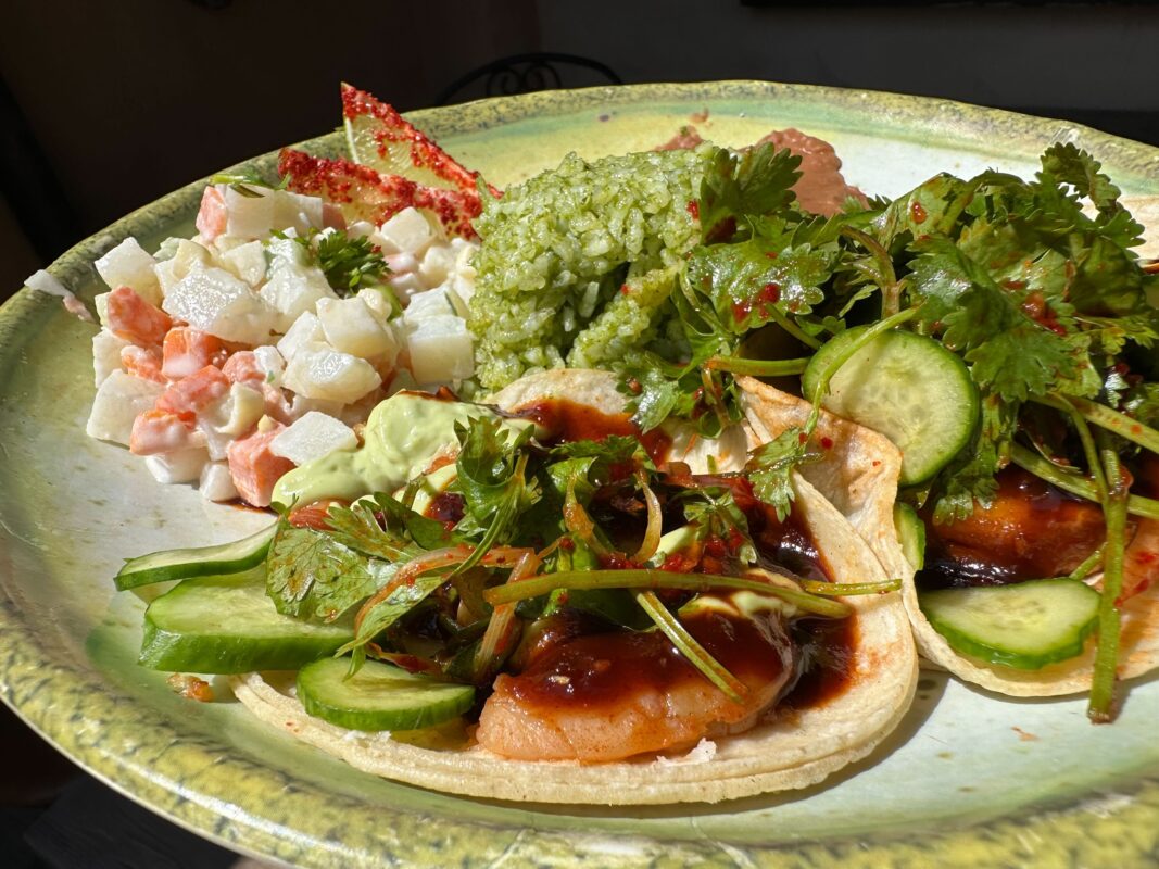 A plate with two tacos topped with lots of greens, alongside a small portion of potato salad and green rice.