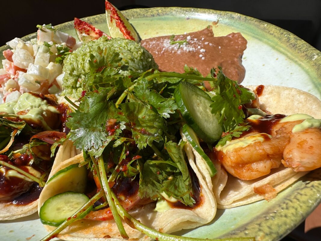 A plate of shrimp tacos topped with cilantro, served with a side of guacamole, beans, and ceviche on a green and yellow plate.