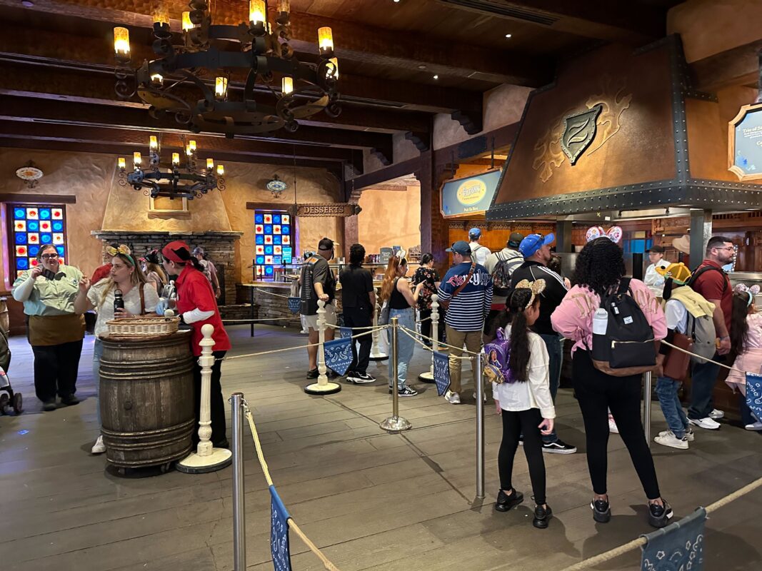 People standing in line inside a themed indoor attraction with rustic decor, stained glass windows, hanging lights, and themed uniforms.