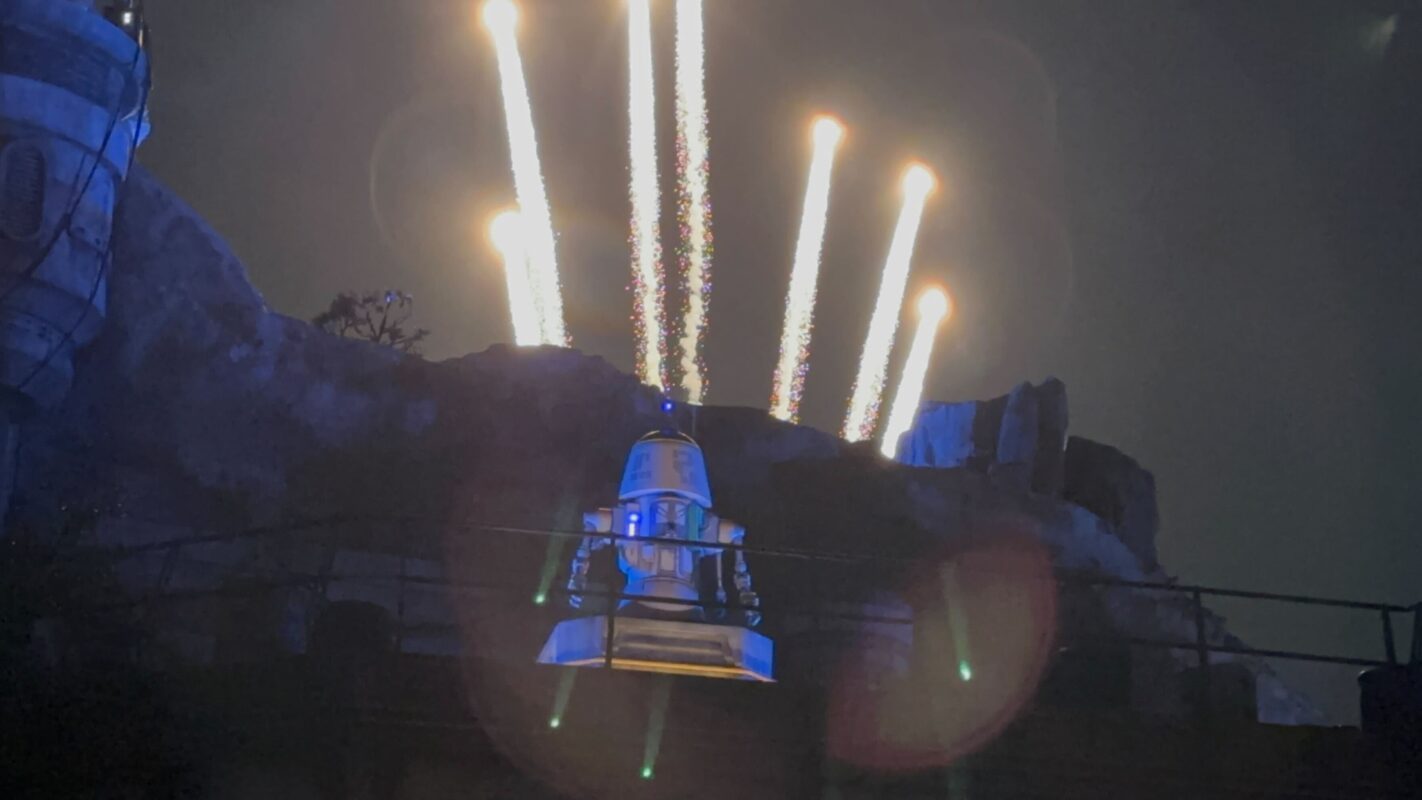 Bard, a droid resembling R2-D2, stands on a platform with several fireworks shooting into the sky in the background.