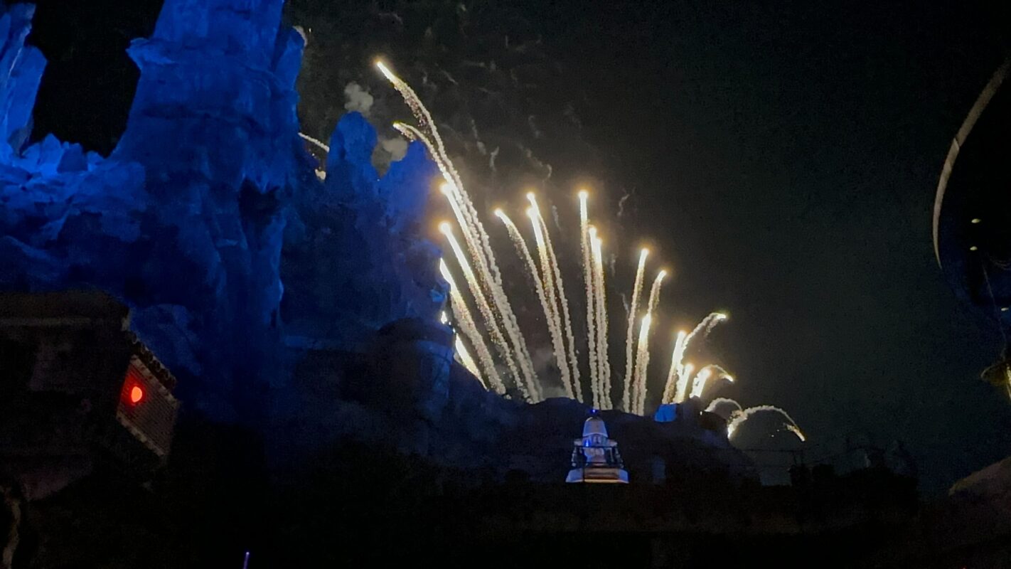 Fireworks light up the night sky against a backdrop of rocky structures during "Fire of the Rising Moons" with a blue hue illuminating part of the scene.