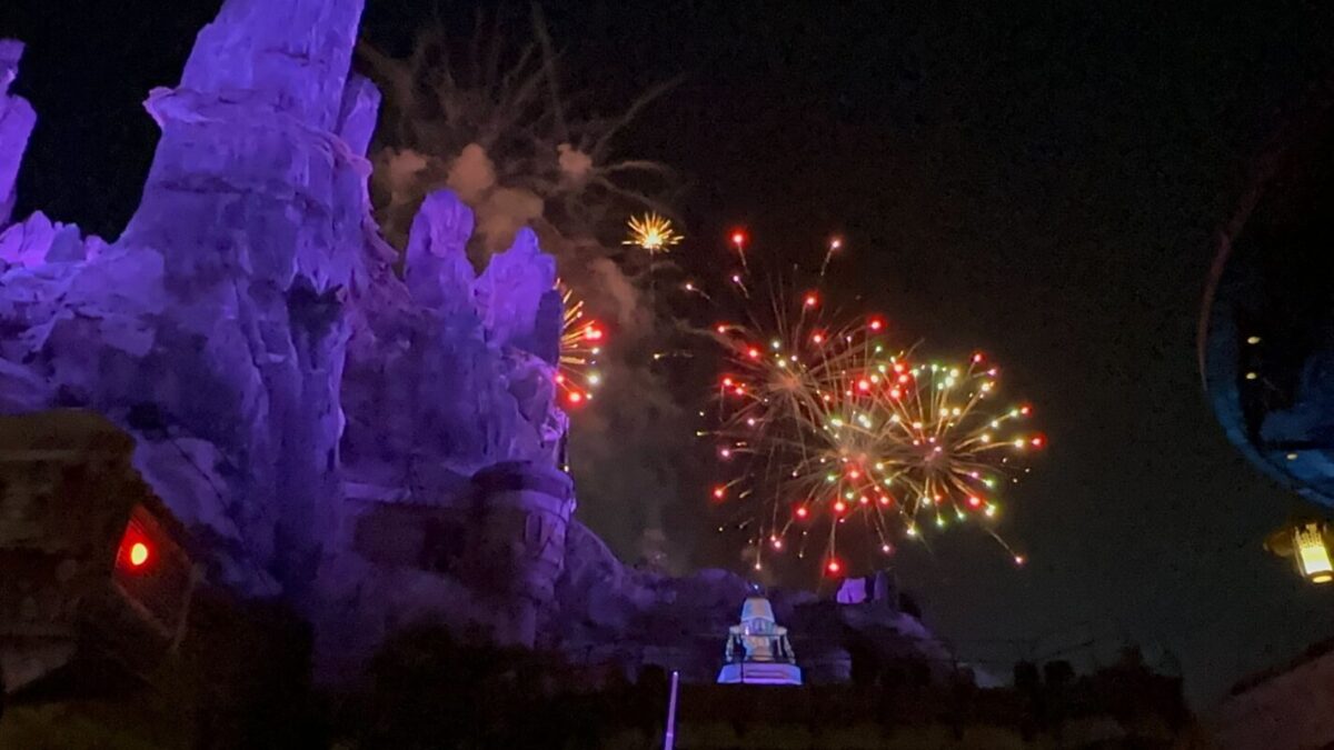 Fireworks burst in the night sky behind a mountain with purple lighting, creating a colorful display of red, yellow, and green lights.