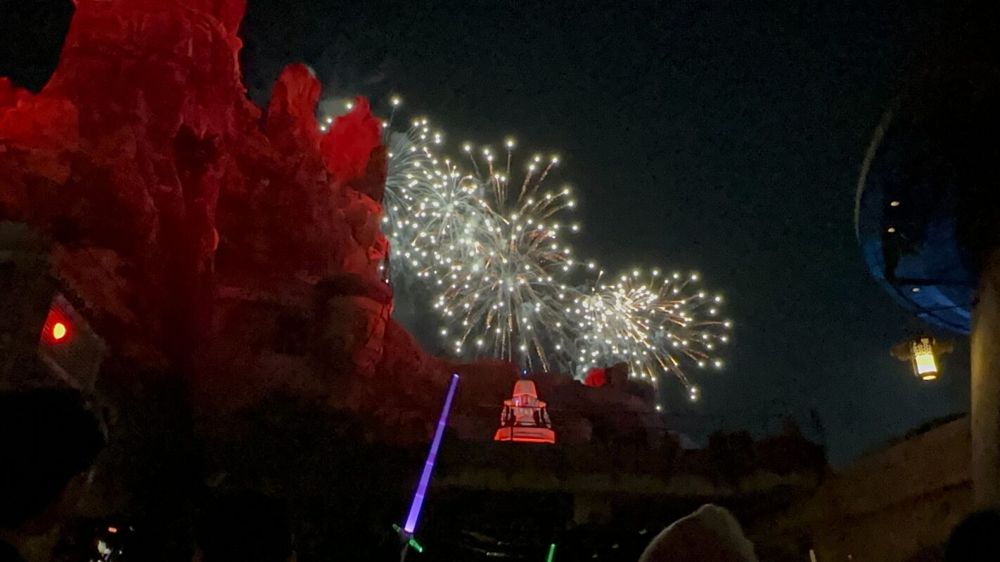 Fireworks light up the night sky above a rocky landscape. Several people holding glowing lightsabers can be seen in the foreground.