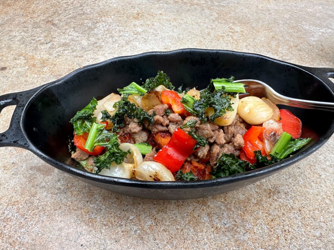 Shaaken Beef with Tatooine Bounty: A skillet containing a colorful stir-fry with ground meat, kale, red bell peppers, and onions, served with a fork on a concrete surface.