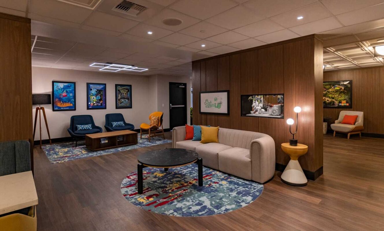 A modern lounge with wooden walls and flooring, featuring a beige sofa, colorful cushions, two blue chairs, a coffee table on a circular rug, wall artwork, and contemporary lighting fixtures.