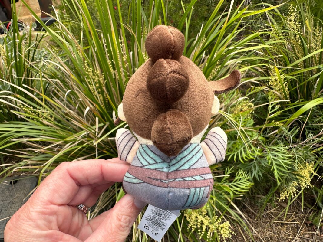 A Wishables plush toy depicting a brown moose wearing a blue and grey striped shirt, held in a hand against a background of green plants.