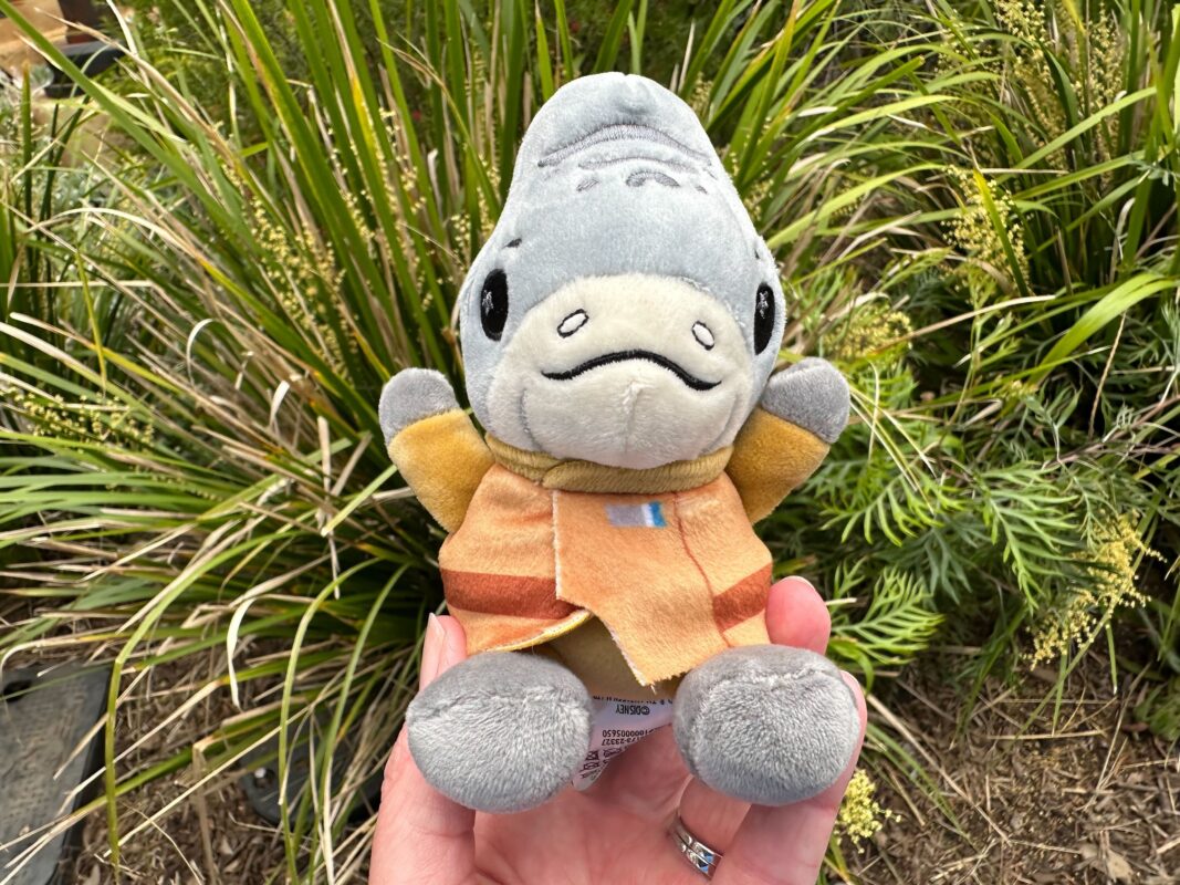 A Wishables plush toy of a smiling shark dressed in an orange and yellow outfit, held in front of greenery.