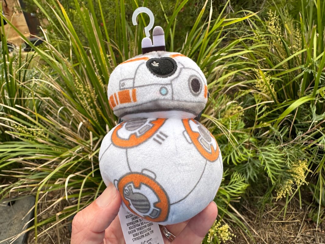 A Wishables plush toy of BB-8 from Star Wars being held in a hand against a backdrop of green shrubs.