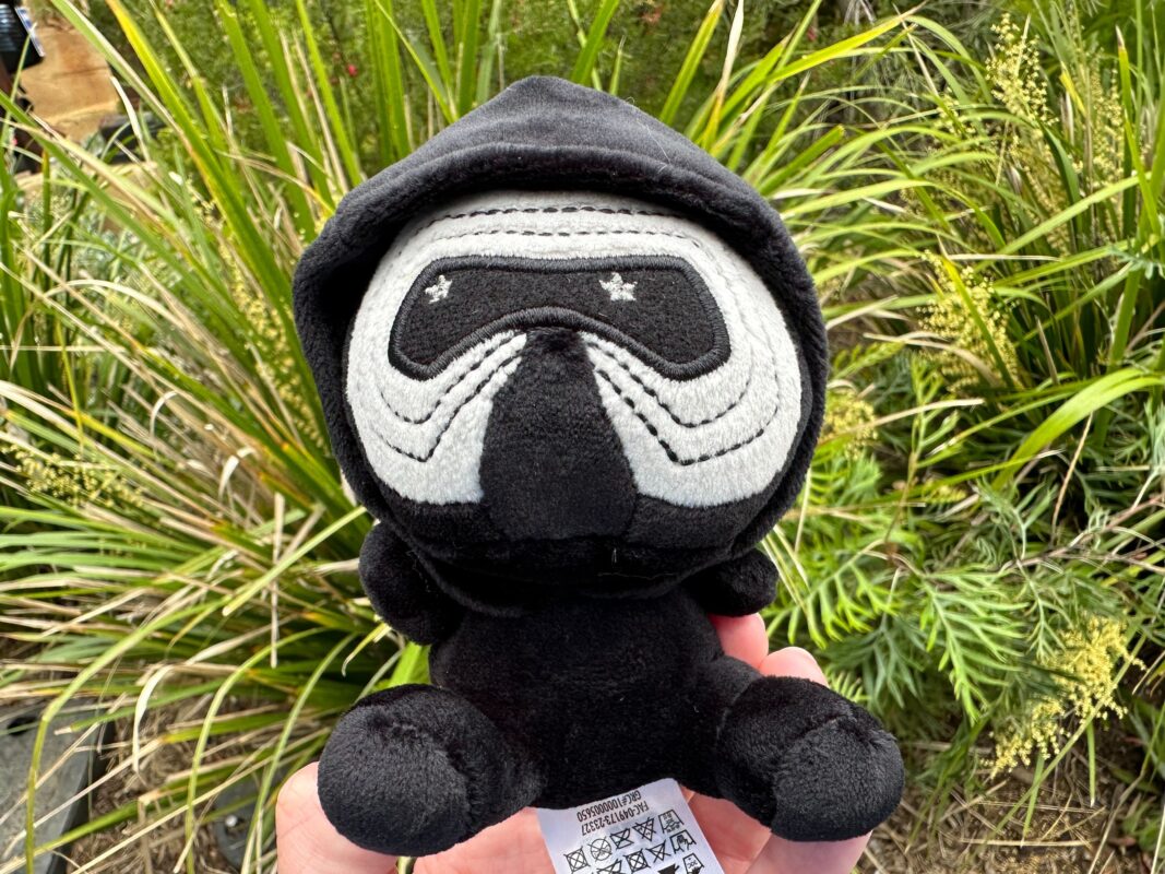 A plush toy resembling a cartoon-like ninja with embroidered eyes, wearing a black hood, held in front of greenery from the Rise of the Resistance.
