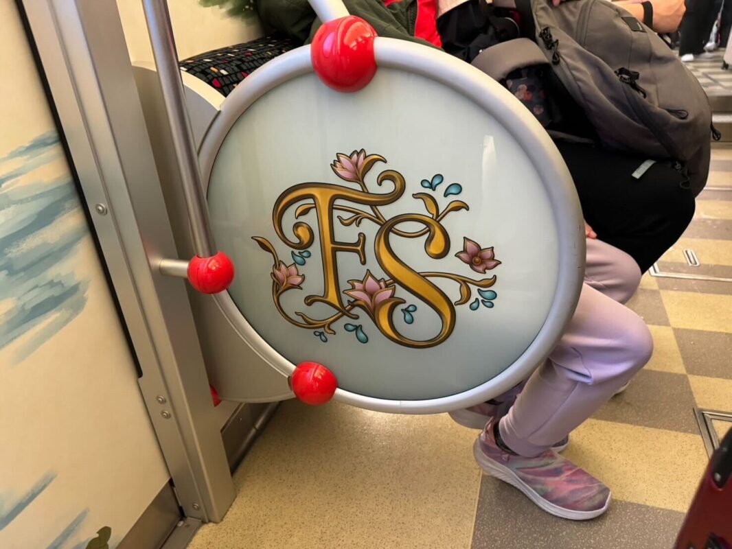 Decorative shield with the initials "fs" mounted on a subway train door, adorned with floral motifs and red orbs, with passengers nearby.