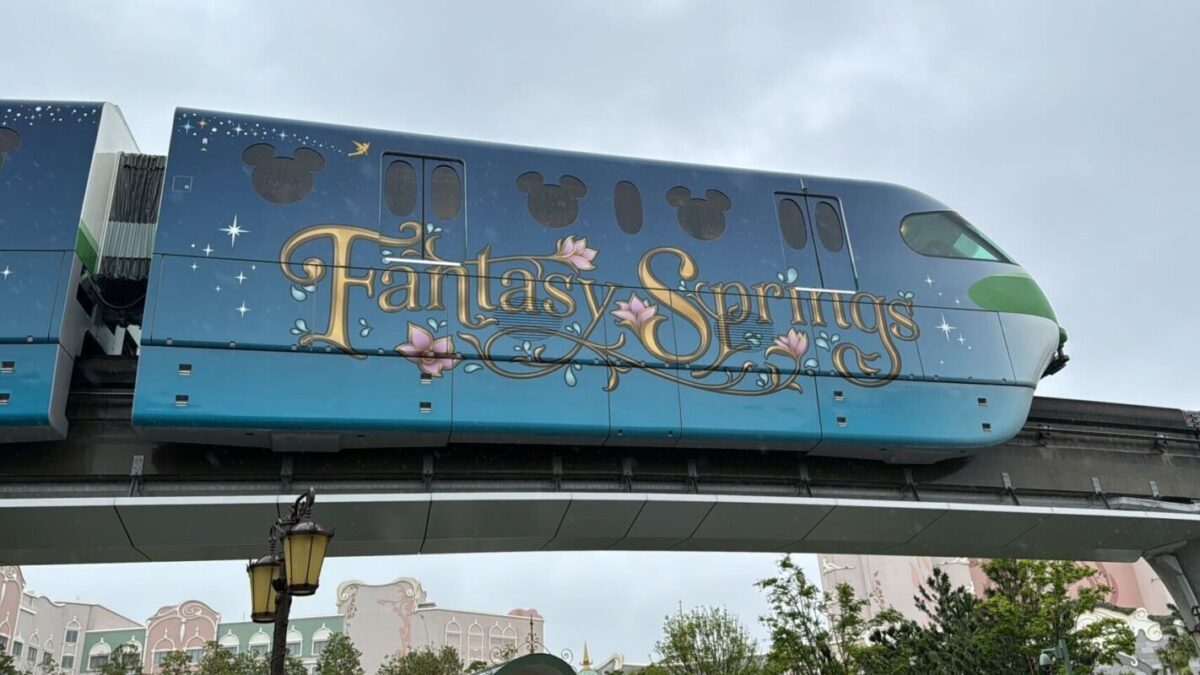 A monorail themed with "fantasy springs" and disney motifs travels above a cityscape on a cloudy day.