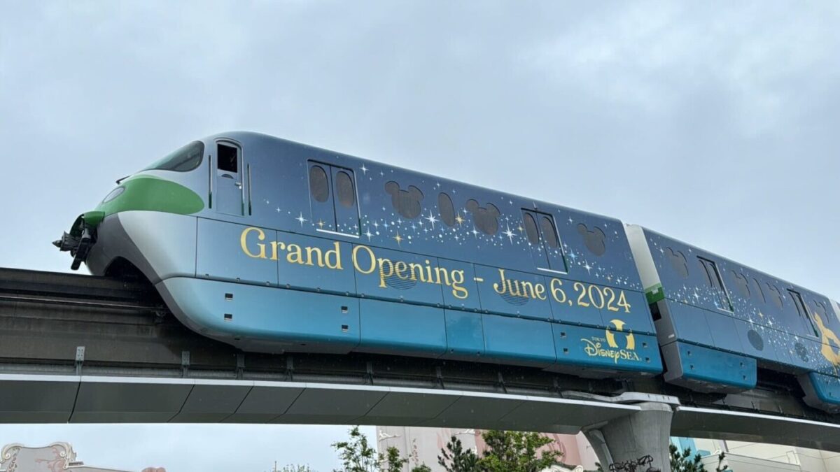 Monorail car with a "grand opening - june 6, 2024" banner and decorative patterns, traveling on an elevated track against a cloudy sky.