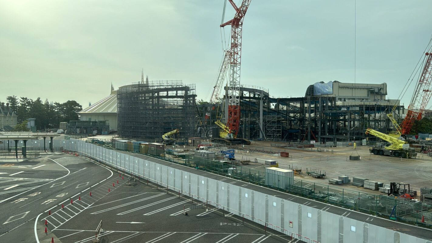 Construction site with steel framework structures, cranes, and machinery. Surrounding the site is a fenced perimeter. The ground shows sections of pavement and open space. The sky is clear with light clouds.