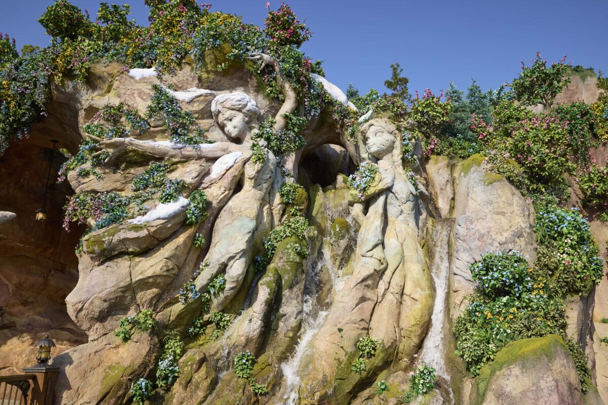 Sculpture of two mythical figures crafted from rock, intertwined with greenery and flowers, with small waterfalls cascading between them under a clear sky.