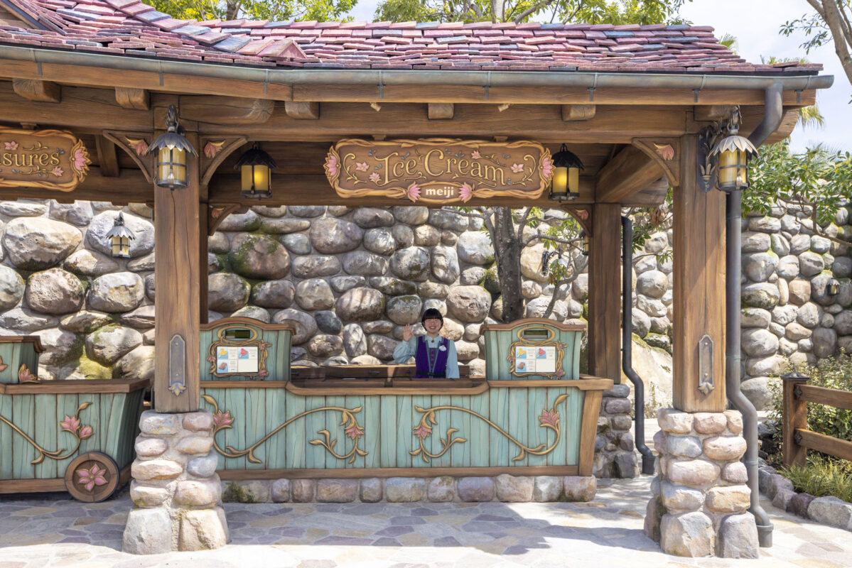 A Cast Member standing at an outdoor ice cream kiosk with a stone facade and a wooden roof, in a sunny park-like setting.