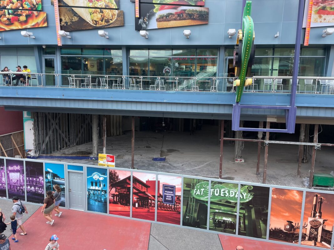 Construction site with a partially demolished building facade, revealing an empty interior. Several colorful posters and signs are displayed on the barriers in front of the site. People walk by.