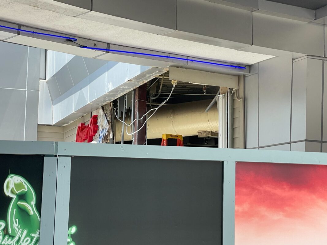 A view of a construction area partially covered by a barricade with a neon parrot sign and a partially visible interior with exposed pipes and ceiling work.