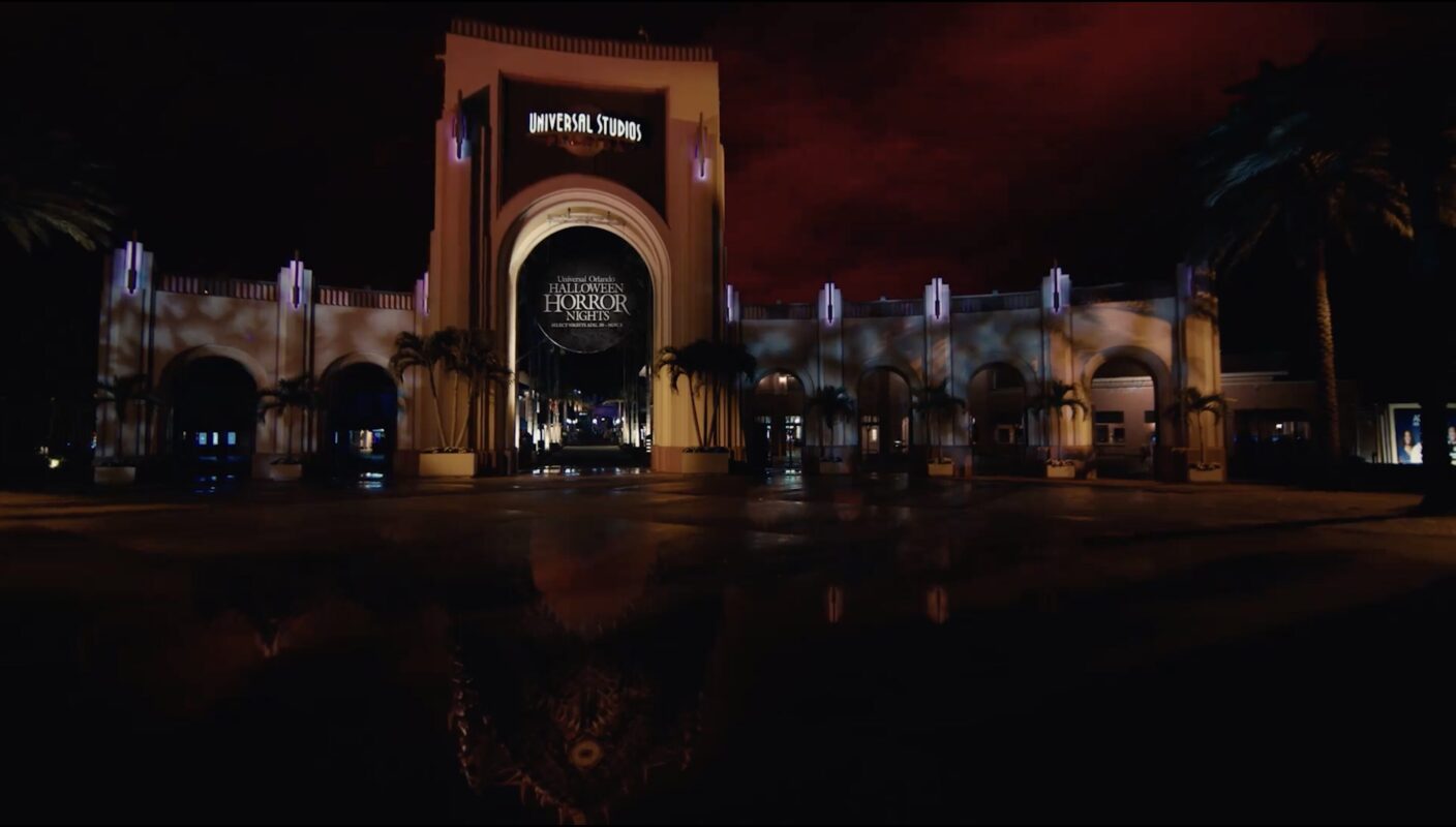 A nighttime view of the illuminated entrance to Universal Studios, decorated for Halloween Horror Nights, with a dark sky and palm trees on each side.