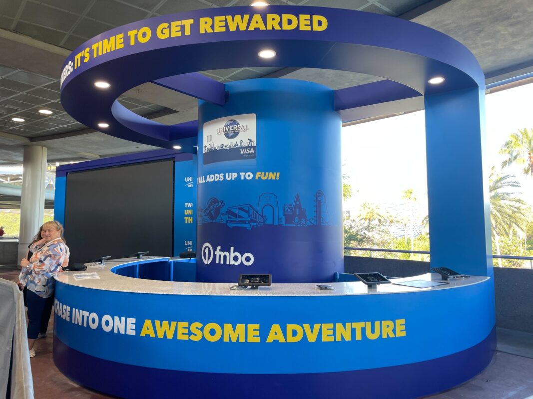 Modern blue information kiosk with promotional FMBO and Universal Visa signage. Counter wraps around a central column. Two people stand near the counter on the left side.