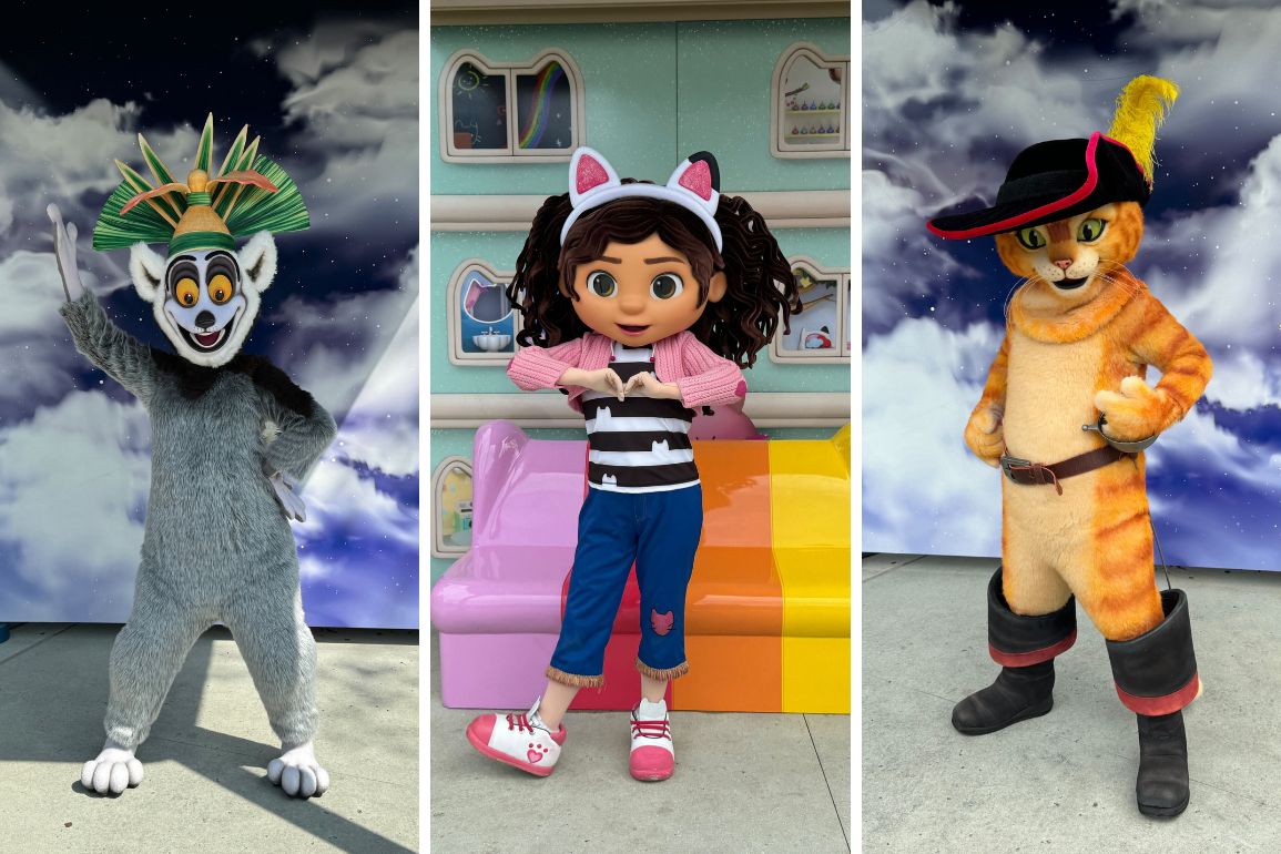Three people in colorful character costumes stand in a row; left to right: a lemur with a crown, a girl with cat ears, and an orange cat with a hat and boots. The backdrop showcases the whimsical DreamWorks Character Zone against a cloudy sky.