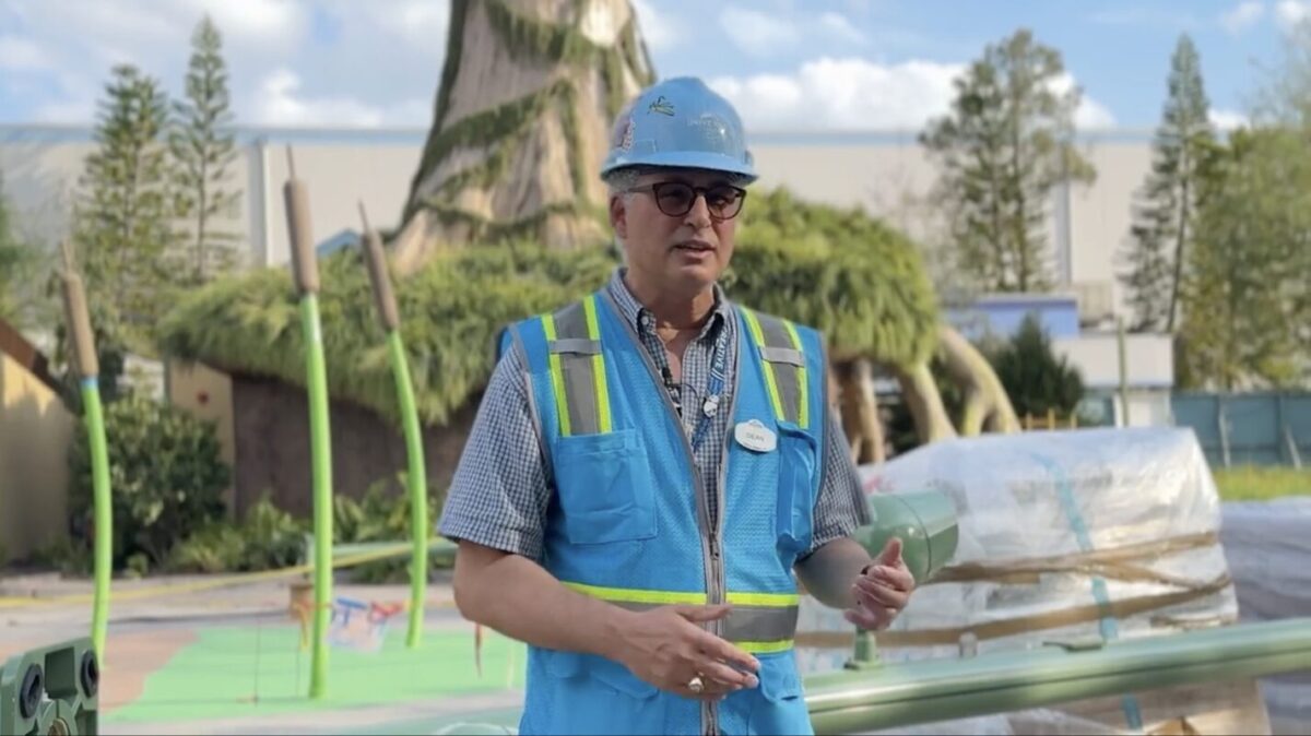 A person wearing a blue construction helmet and a blue safety vest stands on a construction site with green structures and trees in the background.