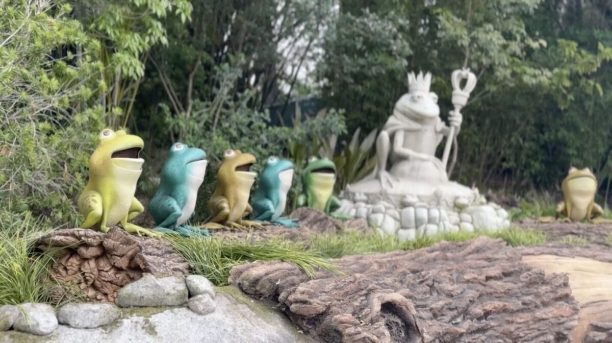 A line of colorful frog statues faces a larger frog statue adorned with a crown and scepter, surrounded by greenery and a rocky foreground.