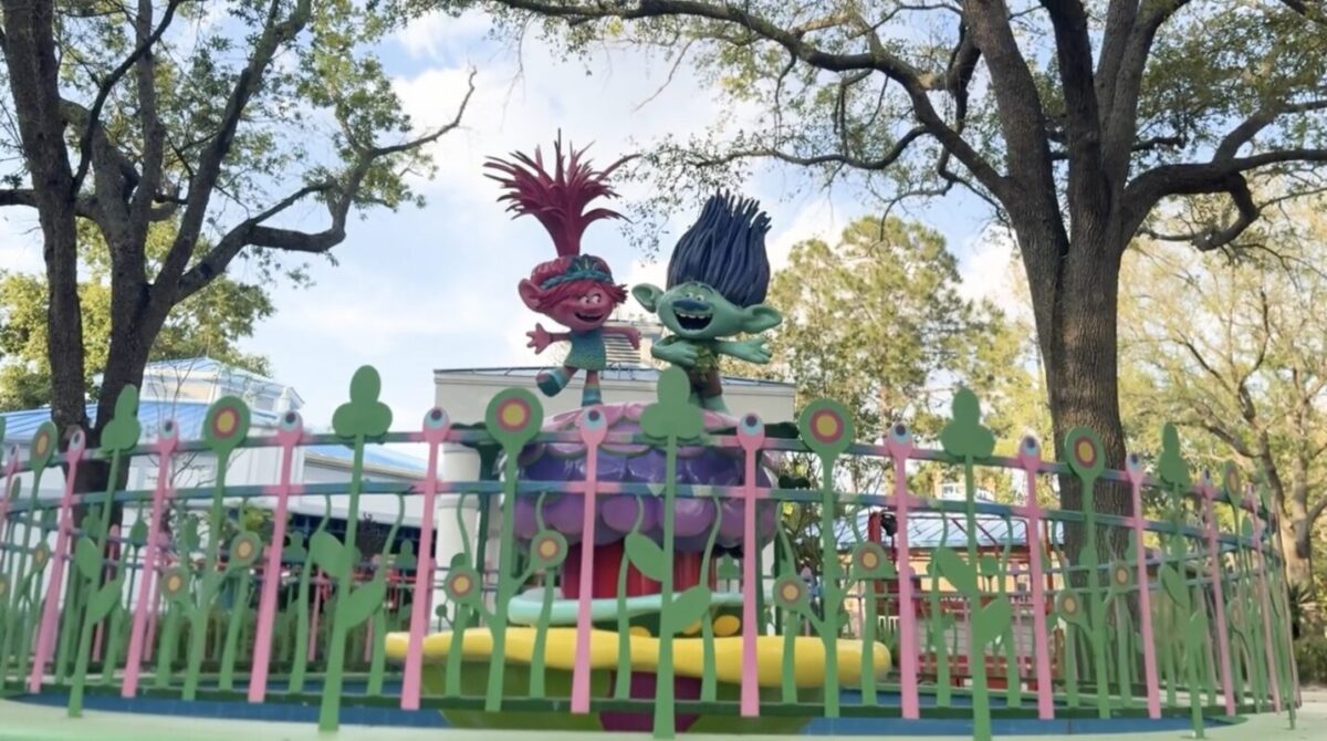 Two colorful troll characters with vibrant hair, one red and one blue, are standing on a vibrant, flower-themed structure surrounded by a decorative fence and trees.