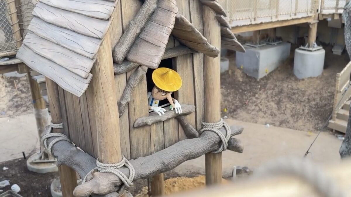 A wooden puppet in a yellow hat peeks out of a window in a rustic treehouse with a wooden roof and ropes. The treehouse is elevated and part of a larger wooden structure.
