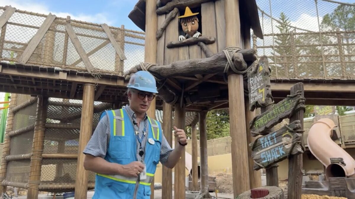 A construction worker in a blue safety vest and hard hat stands near a wooden structure with a figure in a hat peering out from the top. Various signs are visible, including “Ring” and “Hat Shack.”.