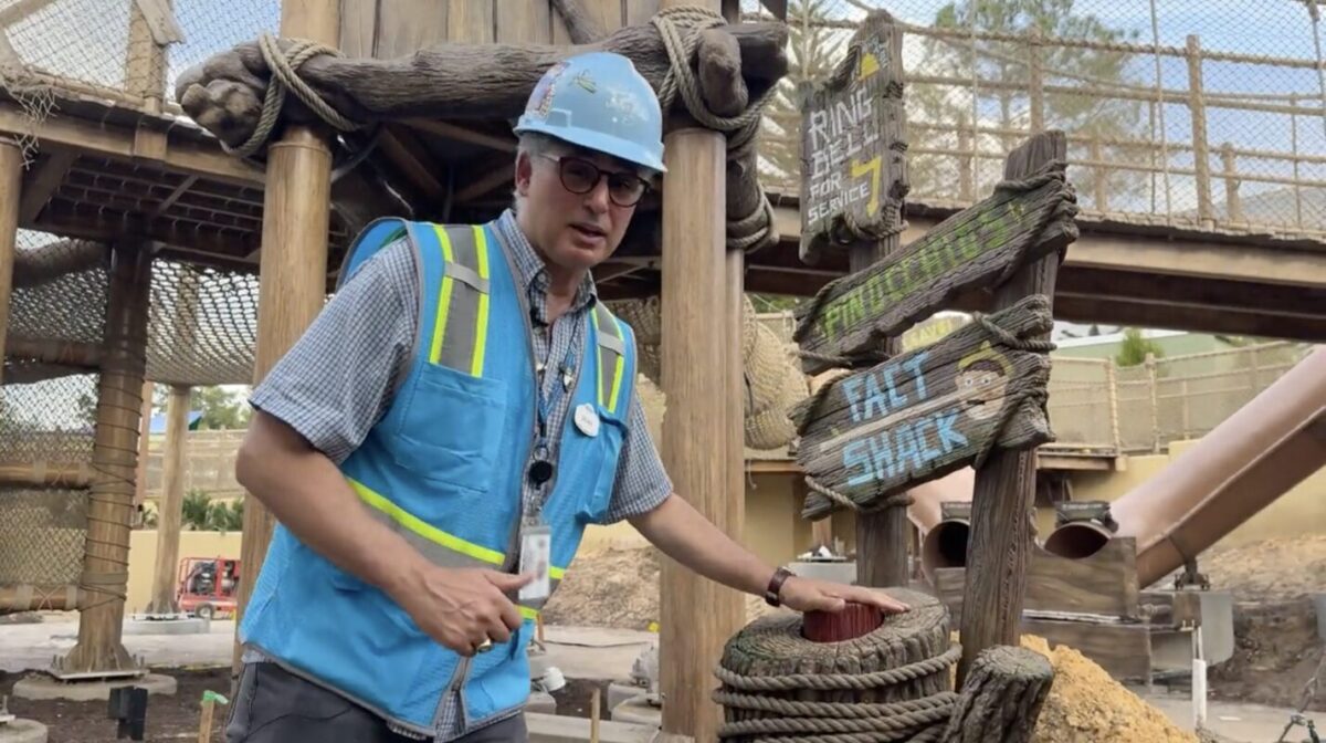 A construction worker wearing a hard hat and blue safety vest stands beside a wooden structure, holding onto a rope and pressing a button. Multiple directional signs are visible in the background.