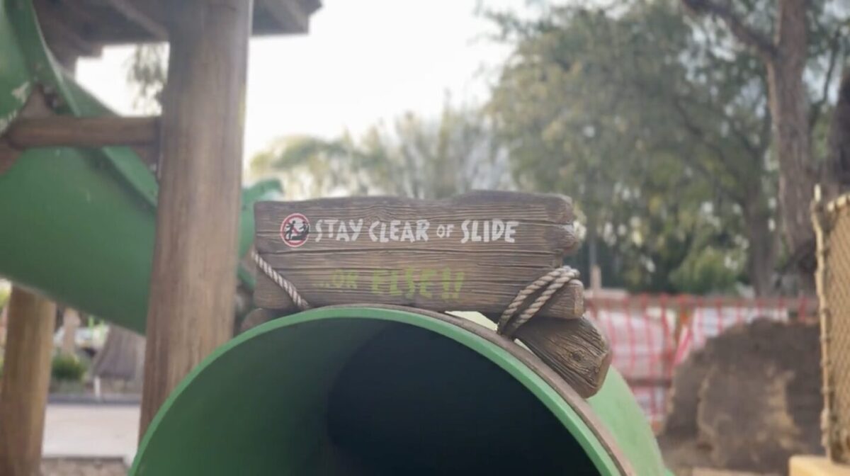 A green playground tube slide with a wooden sign at the top that reads, "STAY CLEAR OF SLIDE BEFORE ENTERING." Trees and blurred structures are visible in the background.