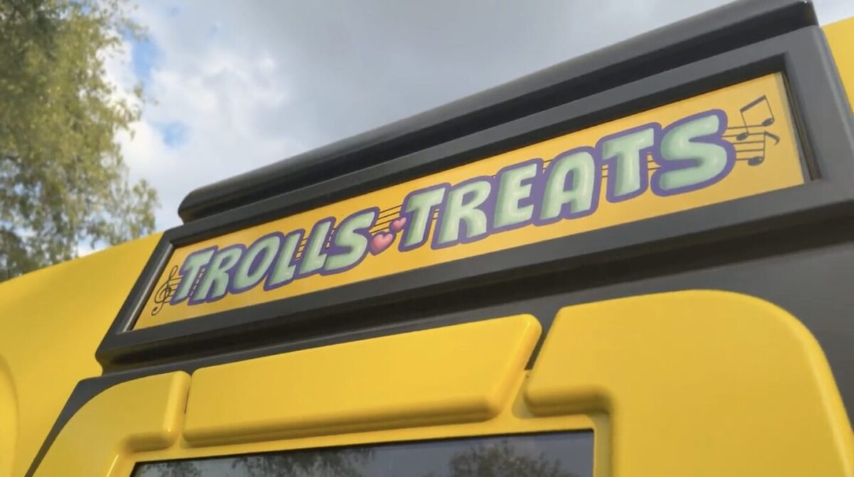 A yellow food truck sign reads "Trolls Treats" against a background of trees and a cloudy sky.