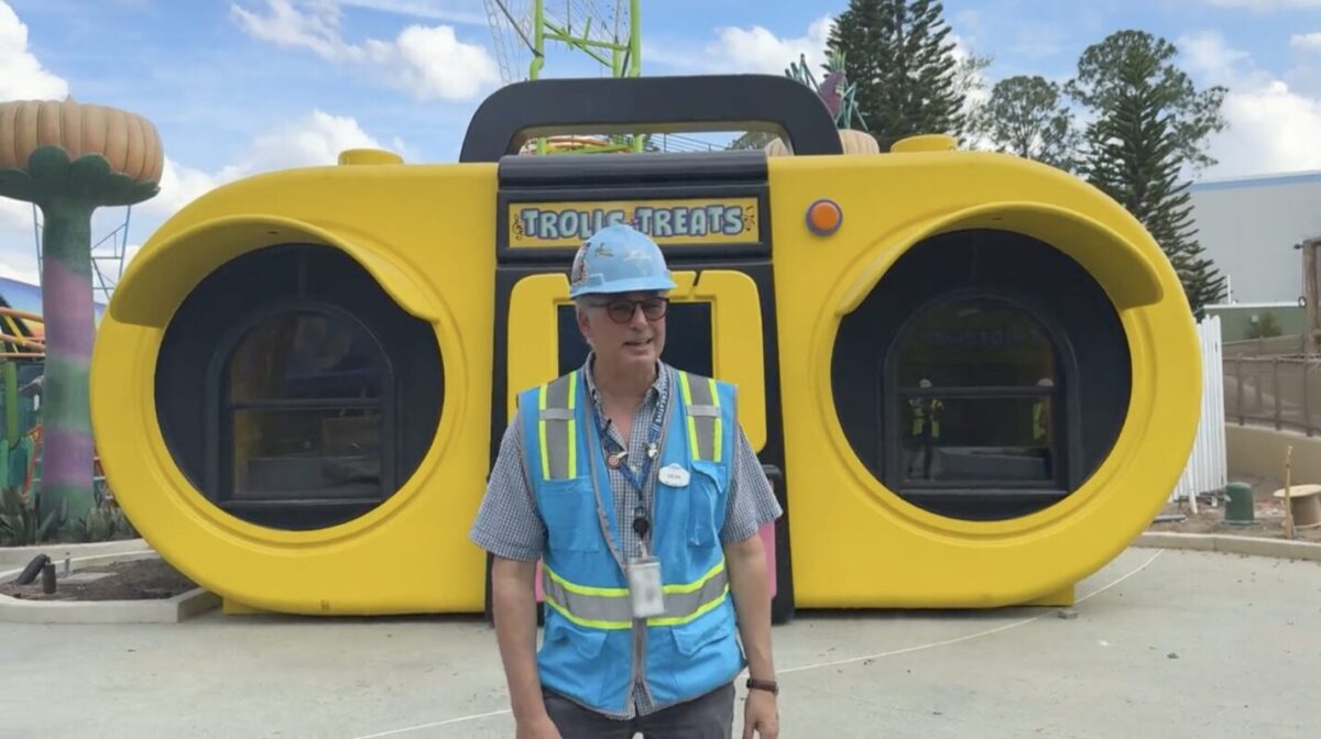A man wearing a blue construction vest and hard hat stands in front of a large, yellow, boombox-themed building at a construction site.