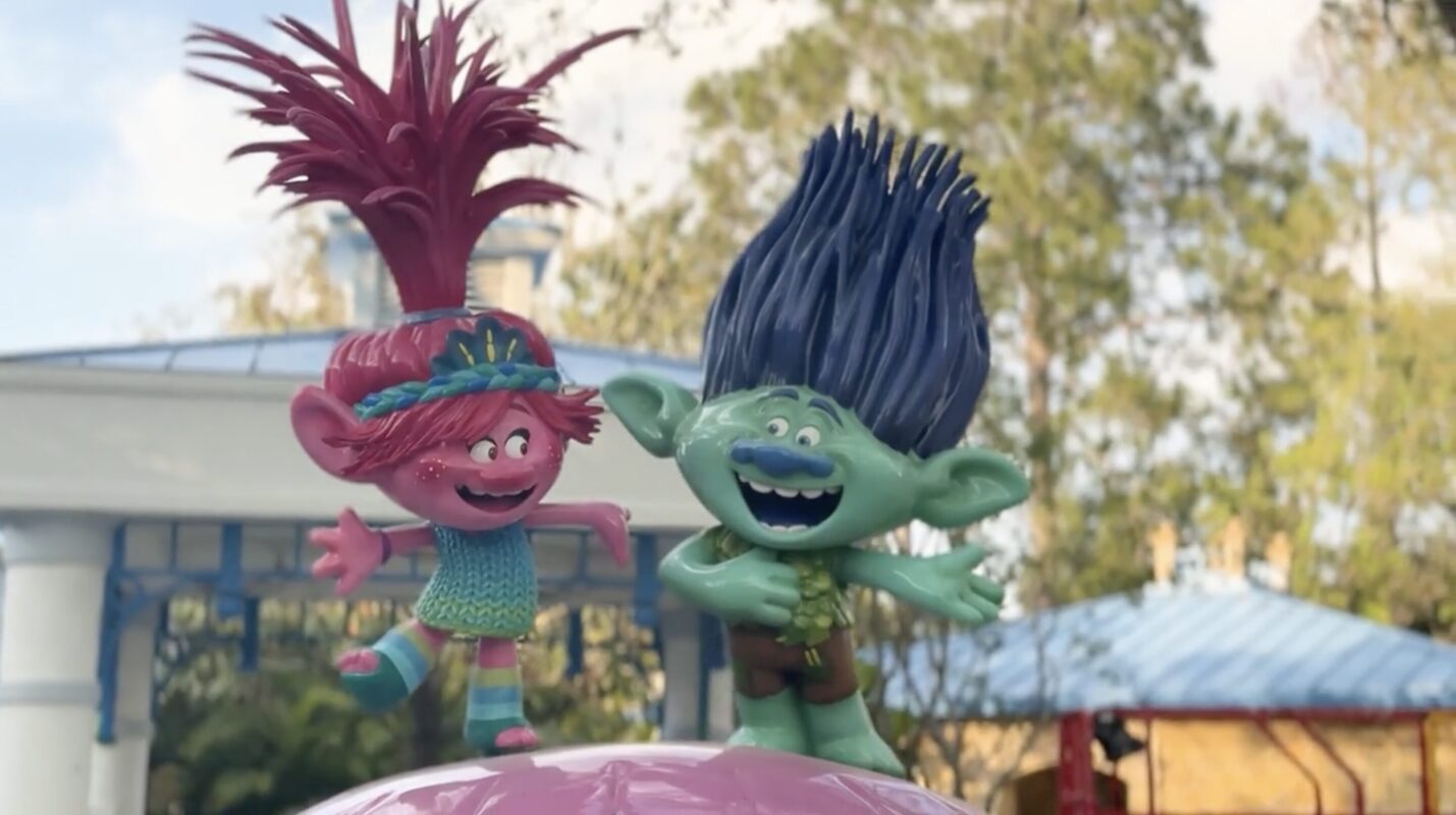 Two colorful troll dolls with exaggerated hair and wide smiles stand joyfully atop a pink surface in an outdoor setting with trees and structures in the background.