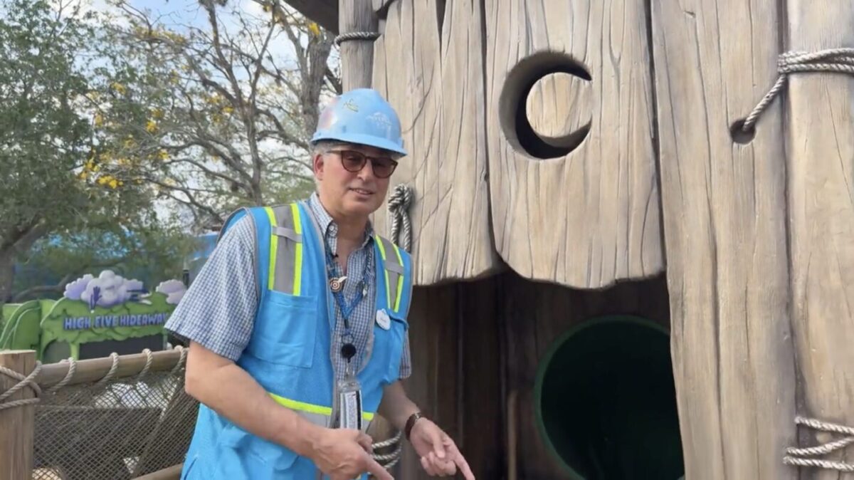 Man wearing a blue safety vest and helmet stands in front of a rustic wooden structure with a circular opening.