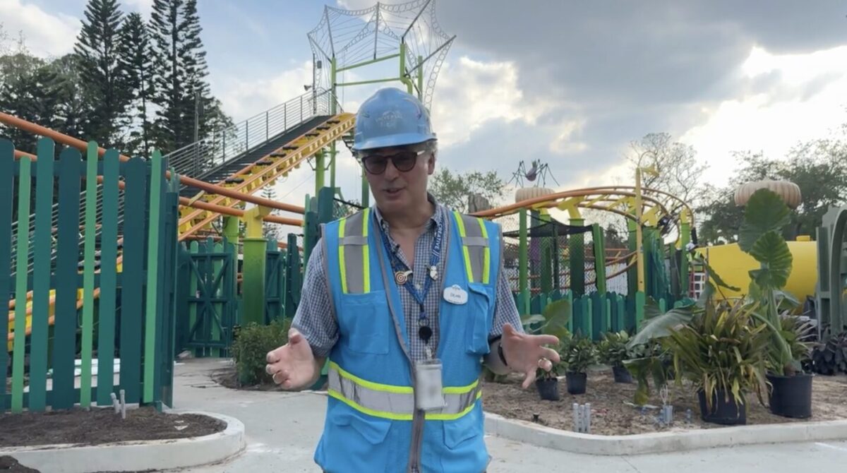 A person wearing a hard hat and reflective vest stands in front of a theme park ride under construction, with plants and a path visible in the foreground.