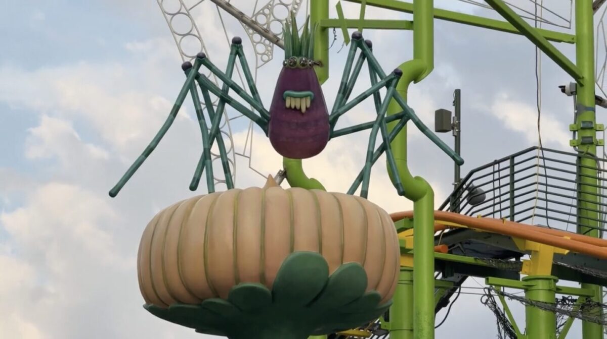 A large mechanical spider-like creature with a purple head and green legs sits atop an oversized orange pumpkin, part of an amusement park ride with green and yellow support structures in the background.