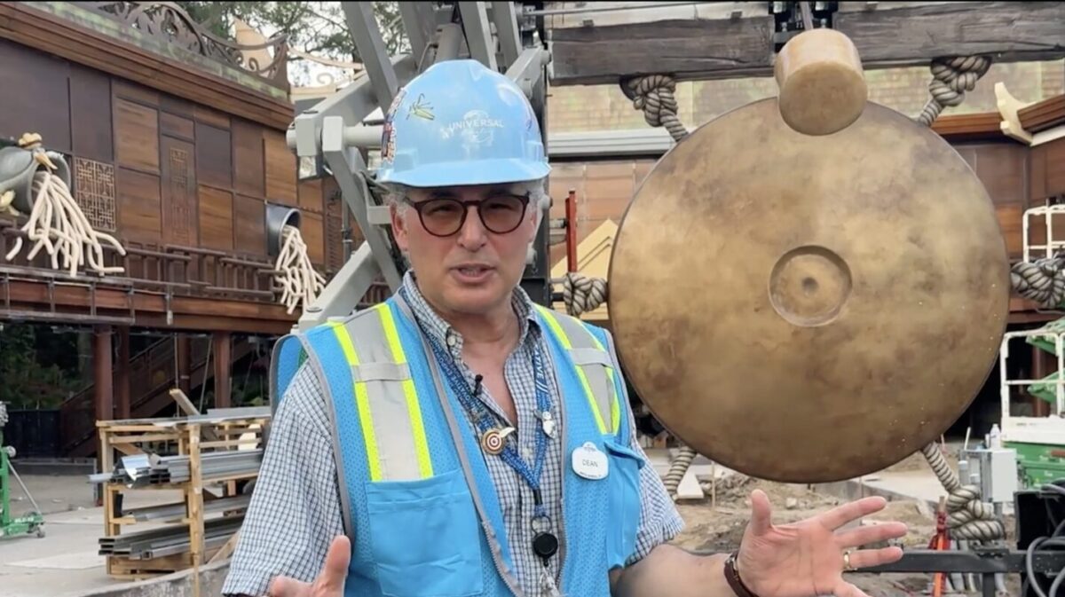 A construction worker wearing a blue hard hat and safety vest stands outdoors near a large gong, surrounded by construction materials.