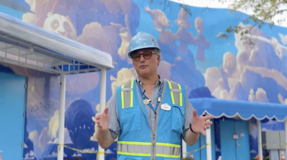A person wearing a blue hard hat, a blue safety vest, and a name badge stands in front of a colorful outdoor mural with clouds. The individual appears to be speaking or gesturing with both hands.