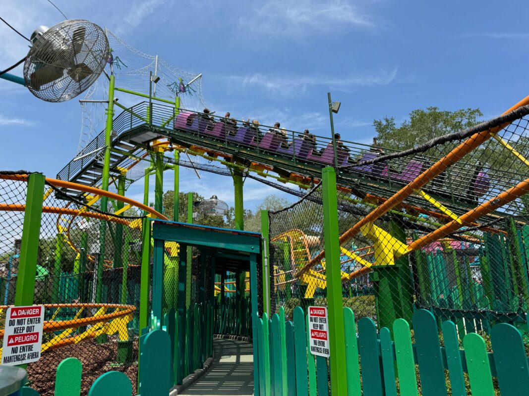 A thrilling trollercoaster ride with people on it is seen in a theme park with vibrant green fencing and safety signs. A large fan is mounted on a pole to the left. Blue sky and trees are in the background.