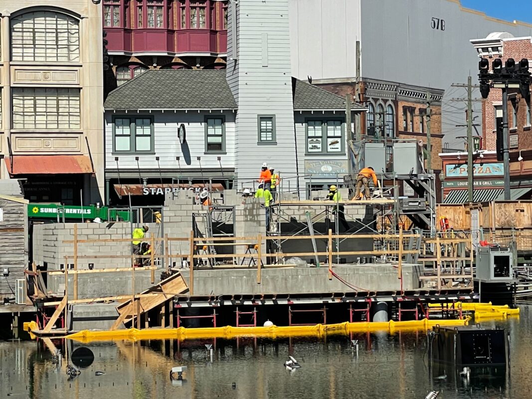 Construction workers building a structure on a site surrounded by water; nearby buildings serve as a backdrop. Yellow flotation barriers are seen in the water.