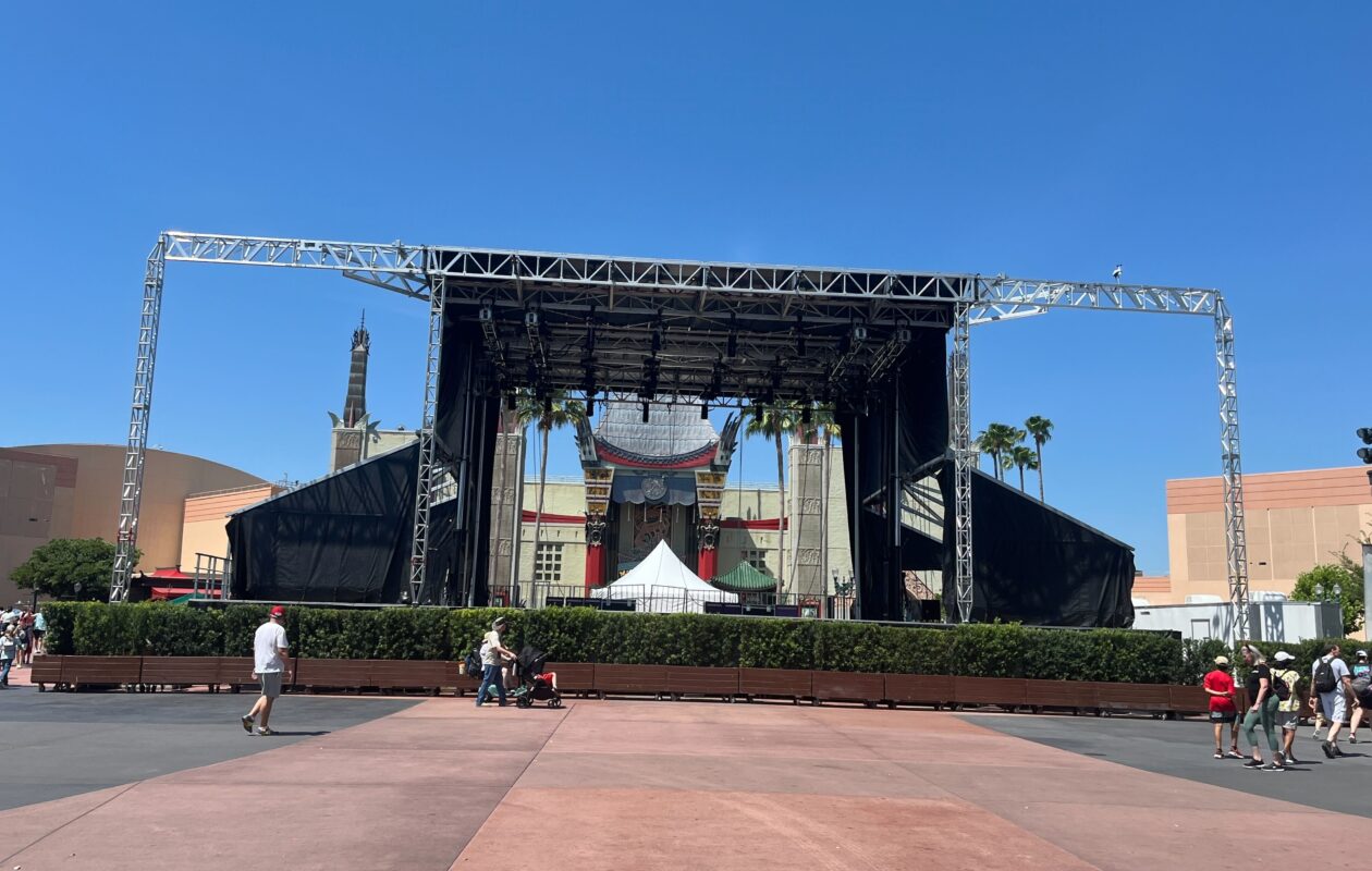 A large outdoor stage setup in front of a replica Chinese theatre with clear blue skies and several people walking nearby, reminiscent of the vibrant atmosphere at Disney's Hollywood Studios.