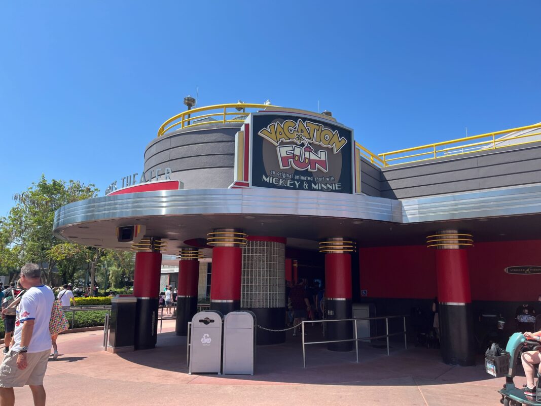 Exterior of the "Vacation Fun" attraction featuring Mickey & Minnie. The building is gray with red columns, a small crowd is gathered outside, and the clear blue sky adds to the daily recap of magical moments shared by visitors.