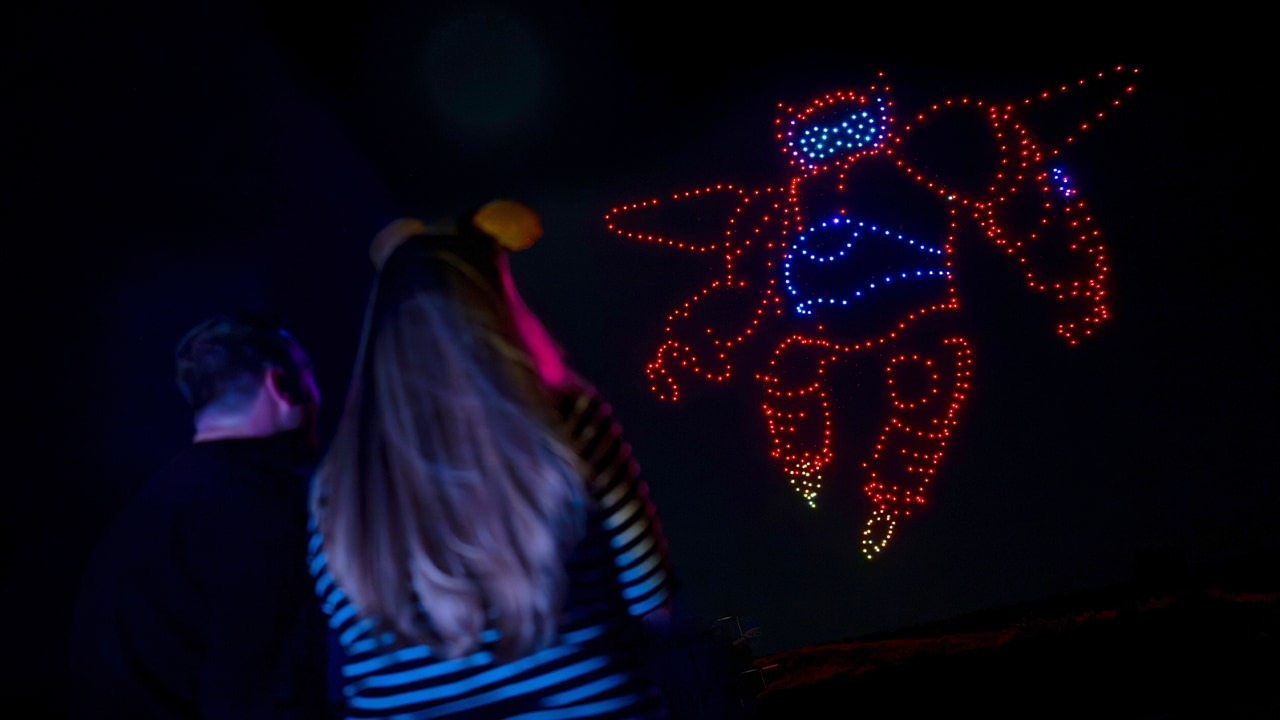 Two people watch a nighttime drone display with red and blue lights forming the shape of Baymax in the sky.